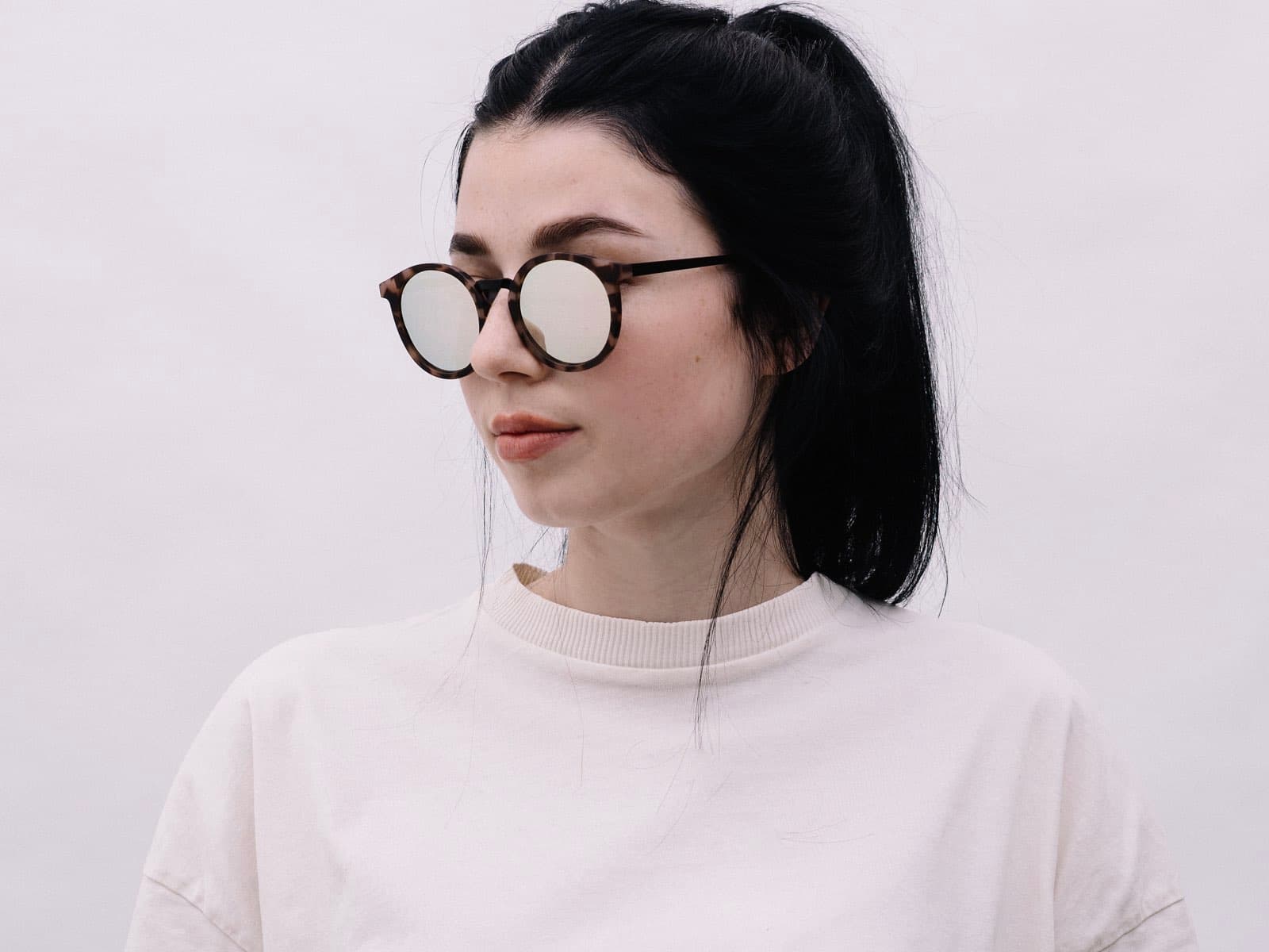 Eine Frau mit schwarzen, hochgebundenen Haare und Brill. Sie trägt ein weißes T-Shirt und hat eine runde Hornbrille auf.